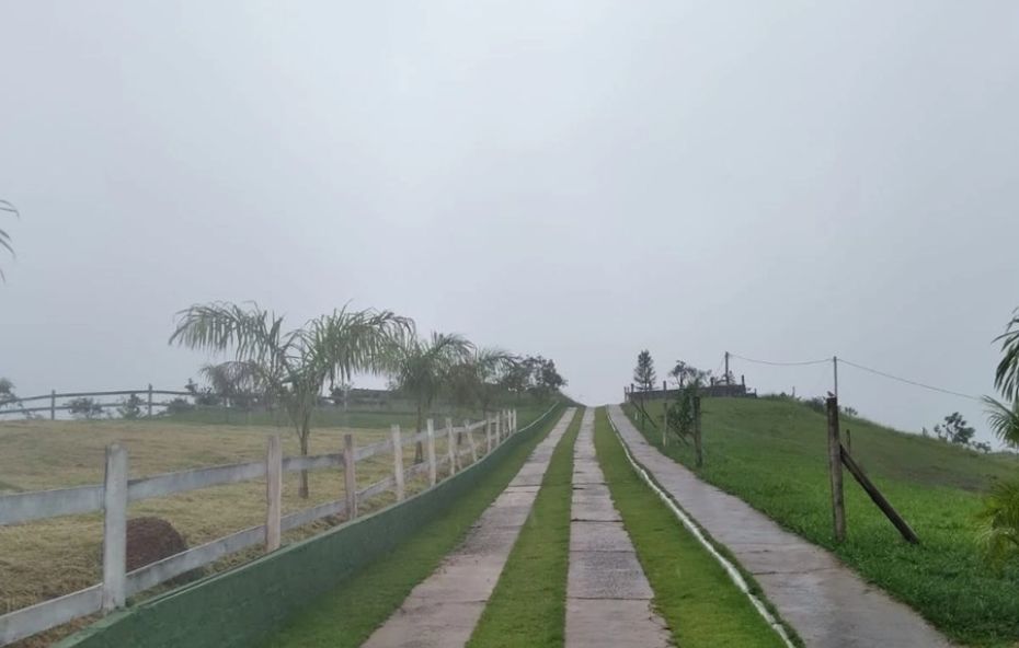 A imagem mostra um amplo terreno de 1.225 metros quadrados cercado por vegetação nativa exuberante no Loteamento Rural Canários da Terra em Ipiabas. O lote apresenta topografia favorável para construção residencial, com pontos de infraestrutura visíveis e uma vista panorâmica para as montanhas preservadas da região serrana.