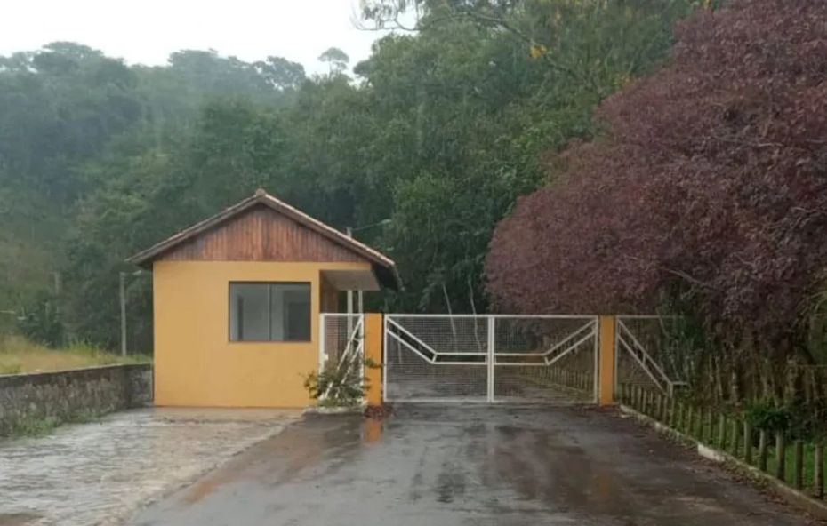 A fotografia exibe um lote plano de 700 metros quadrados dentro do Condomínio Rural Green Village em Ipiabas. O terreno é rodeado por ruas asfaltadas e áreas verdes preservadas, com lagos de pesca esportiva visíveis ao fundo. A vizinhança apresenta casas de alto padrão e uma topografia que favorece a construção imediata com vista panorâmica para as montanhas.