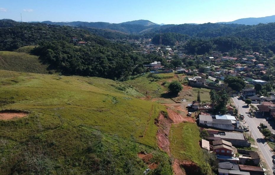A imagem apresenta um terreno de 700 metros quadrados em aclive localizado no Loteamento Senhor do Bonfim em Ipiabas. O lote possui vegetação rasteira e está posicionado estrategicamente em frente ao posto de gasolina, oferecendo uma elevação que garante vista para as montanhas e proximidade com a conveniência urbana local.