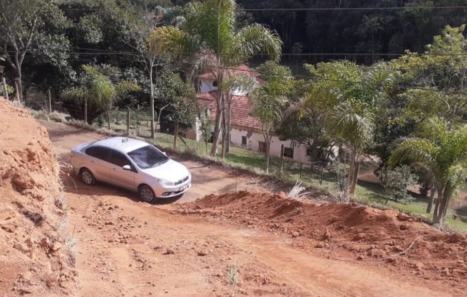 Fotografia em ângulo alto mostra um carro prata em uma estrada de terra íngreme e de terra vermelha. Ao lado, há um barranco de terra cortado. No fundo, abaixo na colina, vê-se uma casa de telhado cerâmico cercada por palmeiras e vegetação densa. A imagem destaca a topografia acidentada do terreno.