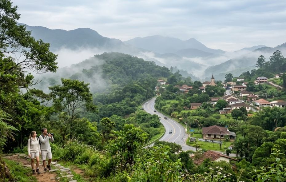 Vista panorâmica de um vale de montanha encoberto por neblina, com um casal de turistas tirando fotos em uma trilha de pedra em primeiro plano. Abaixo, uma estrada sinuosa leva a uma pequena vila aninhada na floresta exuberante.