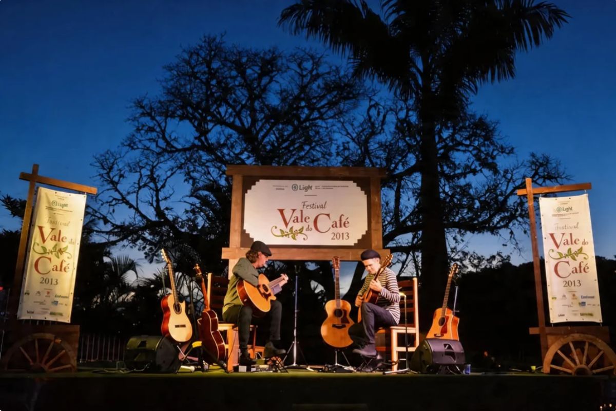Fotografia colorida de dois músicos sentados em um palco iluminado ao ar livre durante o crepúsculo tocando violões acústicos. O palco possui banners laterais e um painel central com o logotipo do Festival Vale do Café de dois mil e treze sob uma árvore de grande porte e um céu azul escuro.