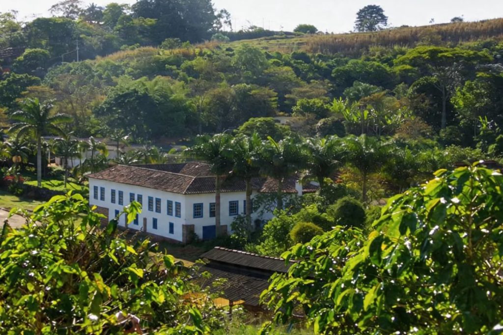 Fotografia colorida em plano aberto mostrando um casarão colonial branco com janelas de molduras azuis aninhado em um vale verdejante com palmeiras imperiais e montanhas densamente arborizadas ao fundo.