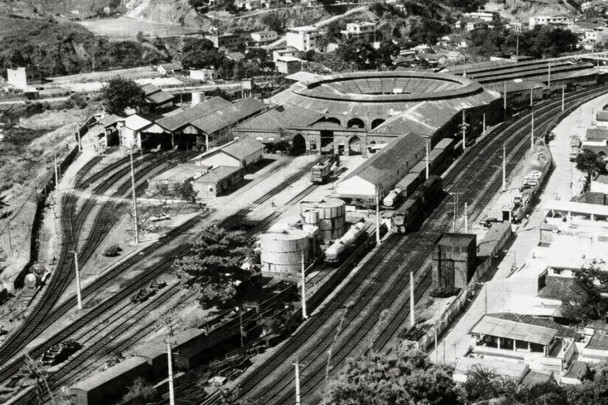 Fotografia aérea em preto e branco mostrando um grande pátio ferroviário com múltiplas linhas de trem paralelas, vagões de carga, tanques de armazenamento e uma grande oficina circular com teto em formato de anel no centro do complexo.