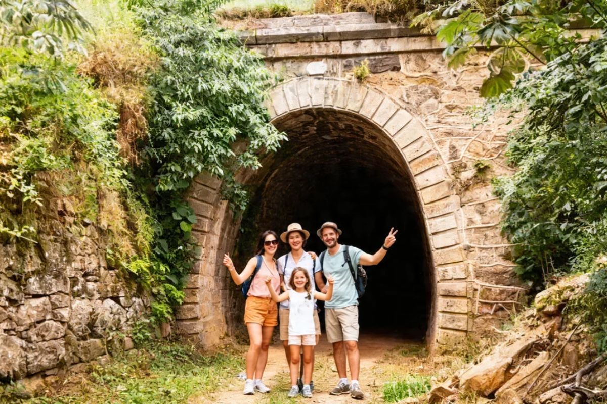 Fotografia colorida em plano médio de quatro turistas sorridentes, dois adultos e duas crianças, posando diante de um grande túnel ferroviário de pedra em Ipiabas. A estrutura do túnel é feita de blocos de pedra rústicos e está coberta por densa vegetação verde nas encostas laterais. A turista à esquerda e as duas crianças fazem sinais de positivo com as mãos enquanto o turista à direita faz o sinal de paz e amor. Todos vestem roupas leves e casuais de verão adequadas para caminhadas ao ar livre. O interior do túnel é escuro e não revela seu fim.