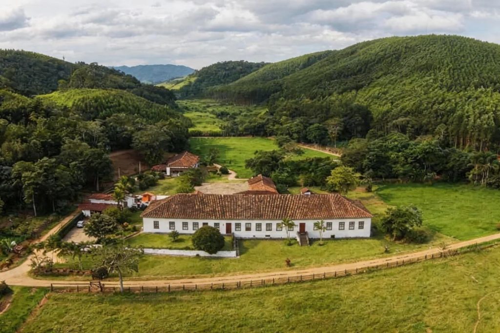 Fotografia aérea colorida de uma fazenda histórica com um longo casarão colonial branco de telhado cerâmico cercado por gramados e montanhas densamente arborizadas sob um céu nublado.