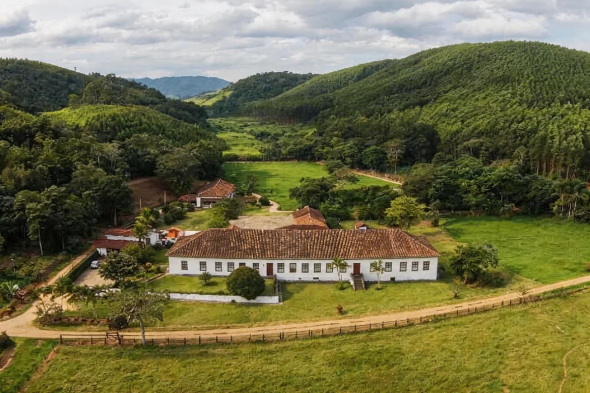 Fotografia aérea colorida de uma fazenda histórica com um longo casarão colonial branco de telhado cerâmico cercado por gramados e montanhas densamente arborizadas sob um céu nublado.