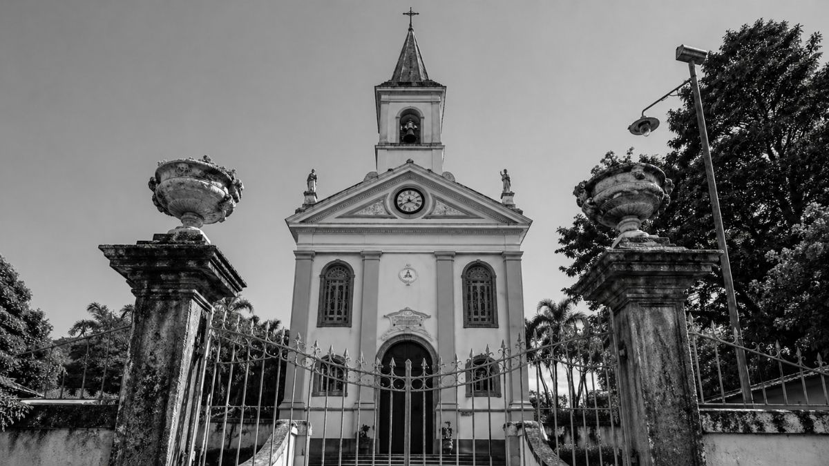 Fotografia em preto e branco da fachada frontal da Catedral de Sant'Ana vista através de um portão de ferro trabalhado.
