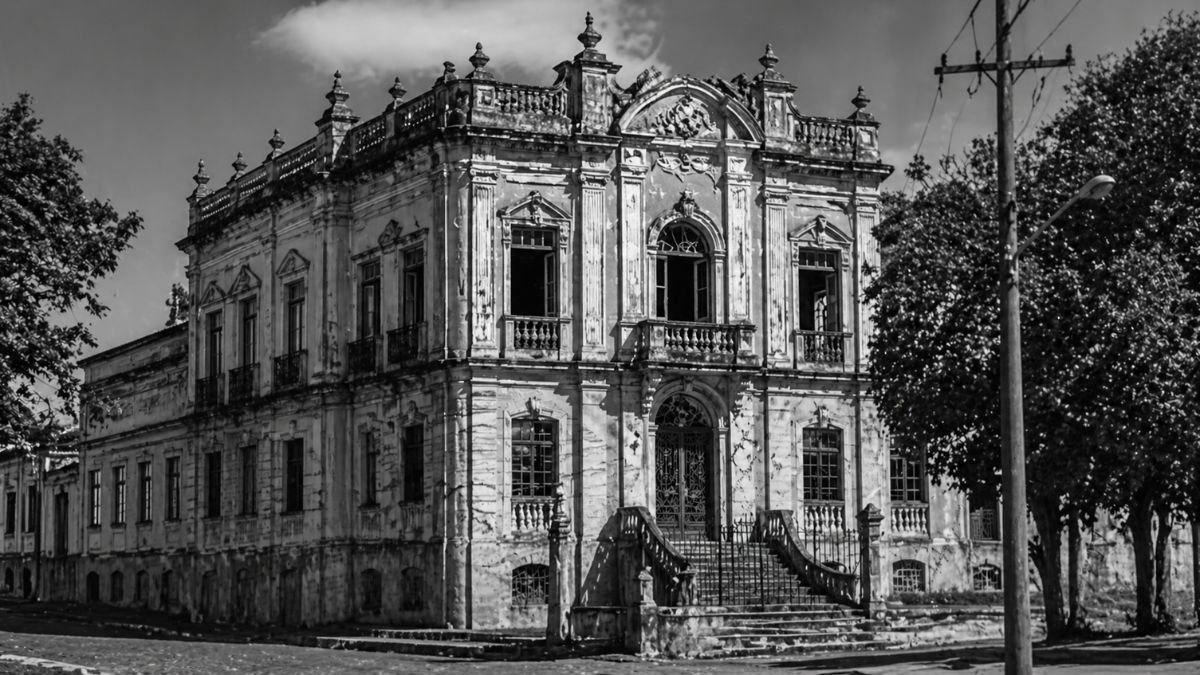 Fotografia em escala de cinza de um imponente casarão histórico com arquitetura clássica, exibindo detalhes em relevo, grandes janelas e escadaria central.