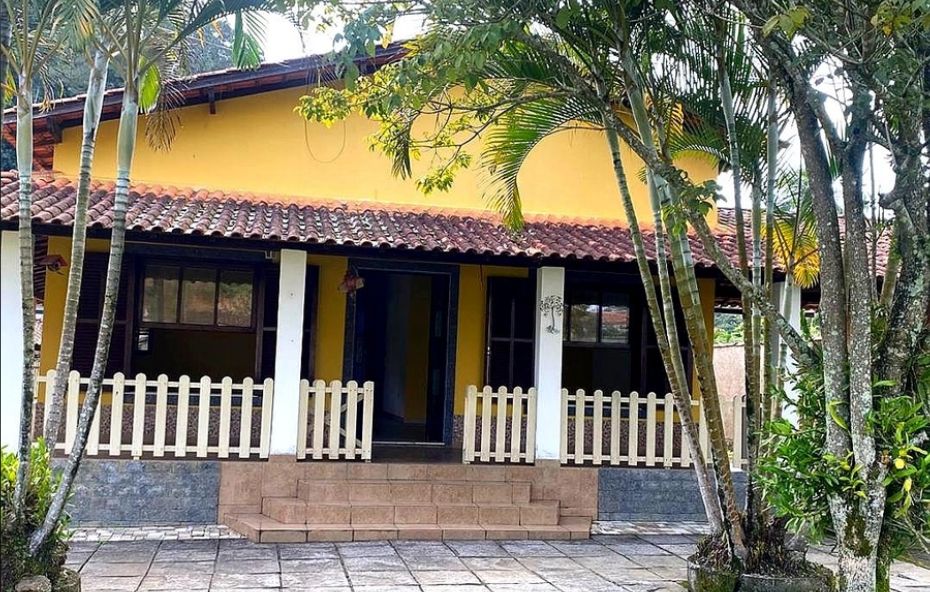 Fachada de casa amarela com varanda protegida por pequena cerca branca e cercada por árvores altas e jardim bem cuidado.