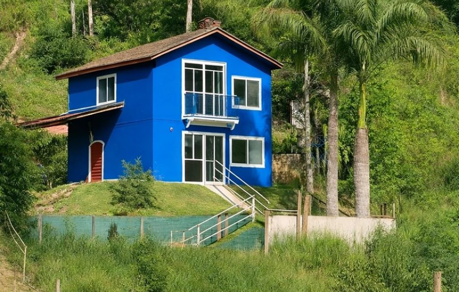 Casa de dois pavimentos pintada em azul vibrante com detalhes brancos situada em uma encosta gramada próxima a palmeiras altas.
