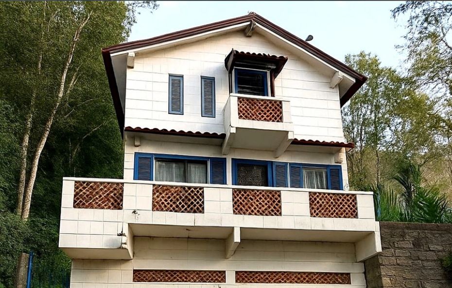 Fachada de uma casa de dois pavimentos em estilo chalé com paredes brancas e janelas azuis destacando varandas com detalhes em treliça cerâmica.
