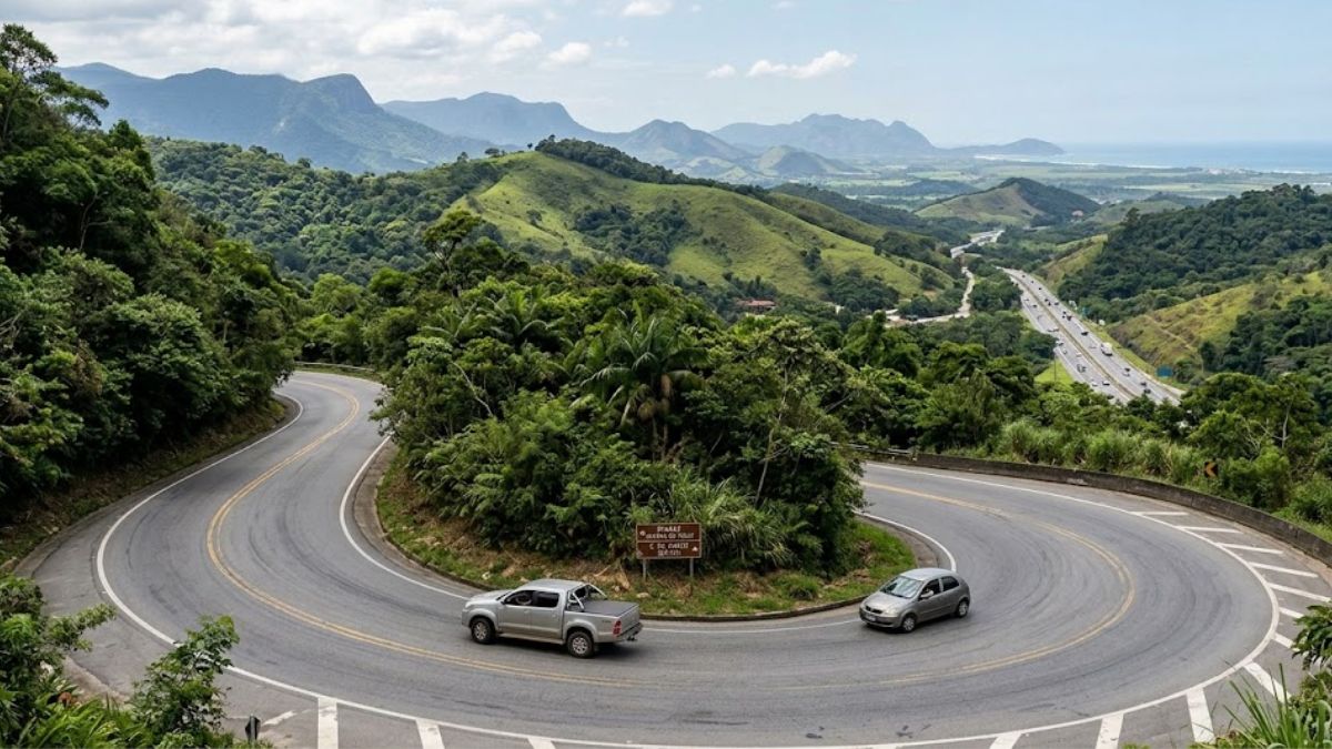 Uma fotografia de ângulo elevado de uma estrada de montanha pavimentada de pista dupla com uma curva em forma de U acentuada, cercada por floresta densa e com vista para uma costa e montanhas distantes, apresentando dois carros prateados.