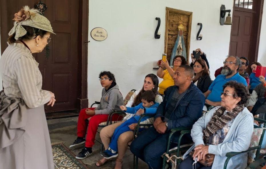 Fotografia colorida capturada em um ângulo médio sob a luz do dia no interior de uma construção histórica, possivelmente uma fazenda. À esquerda, uma mulher idosa de perfil, vestindo um traje completo de época com blusa listrada e saia longa bege, além de um chapéu decorado, está em pé sobre um tapete estampado. Ela fala para um público de cerca de dez pessoas sentadas em cadeiras de ferro esverdeadas rústicas dispostas em fileiras no lado direito. O público, composto por adultos e crianças de diversas idades, está vestido com roupas casuais contemporâneas e presta atenção. Um jovem de calças vermelhas e casaco cinza está sentado na primeira fileira e olha para a oradora. Uma mulher e um homem mais idoso estão sentados na fileira de trás, ambos olhando para a oradora. Outros membros do público mais ao fundo também estão atentos, alguns usando celulares para registrar a cena. Ao fundo, uma parede branca com uma placa dourada com texto ilegível, duas arandelas de ferro preto com velas decorativas, um nicho com uma pequena estátua da Virgem Maria e um sino de bronze pendurado completam o cenário histórico e rústico.