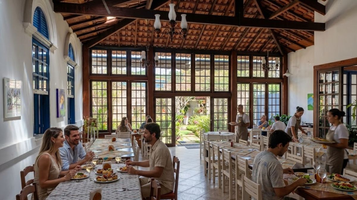 Um restaurante acolhedor com telhado de madeira e grandes janelas abertas para um jardim está movimentado com casais e famílias almoçando e garçons servindo pratos típicos.