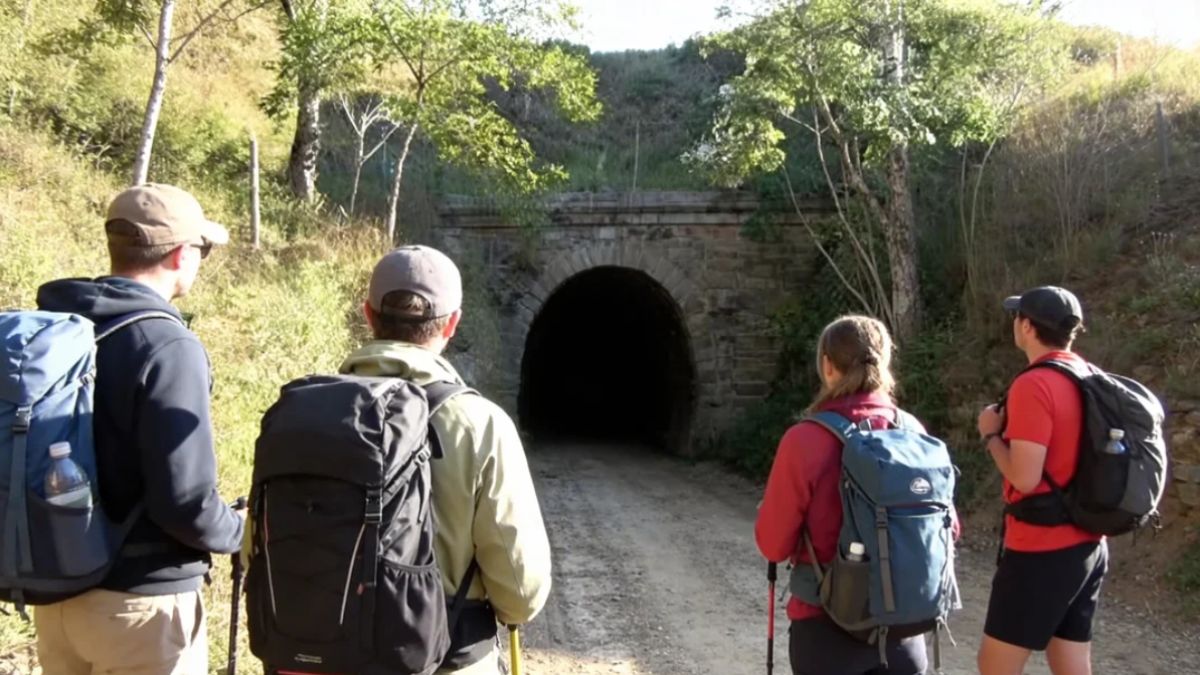 Quatro trilheiros com mochilas cargueiras e bastões de caminhada aparecem de costas em uma trilha de terra batida observando a entrada monumental de pedra do Túnel Velho de Ipiabas que está cercada por encostas gramadas e árvores sob luz solar intensa.