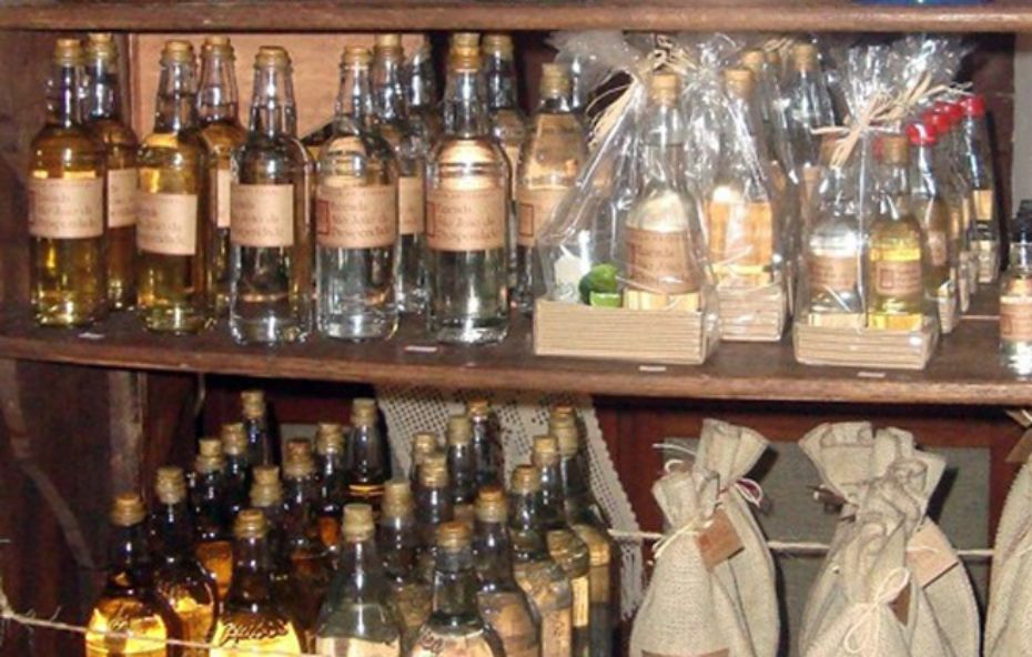 Fotografia colorida em close de prateleiras de madeira rústica repletas de produtos artesanais. Na prateleira superior, fileiras de garrafas de vidro transparentes e âmbar contêm cachaça artesanal com rótulos de papel pardo. Algumas garrafas estão embaladas individualmente em sacos plásticos transparentes com laços de palha, acompanhadas de pequenos brindes em caixas de papelão. Na prateleira inferior, mais garrafas de cachaça estão organizadas ao lado de sacos de estopa amarrados com cordão, possivelmente contendo grãos de café. A iluminação é quente e destaca os tons amadeirados e o brilho do vidro.