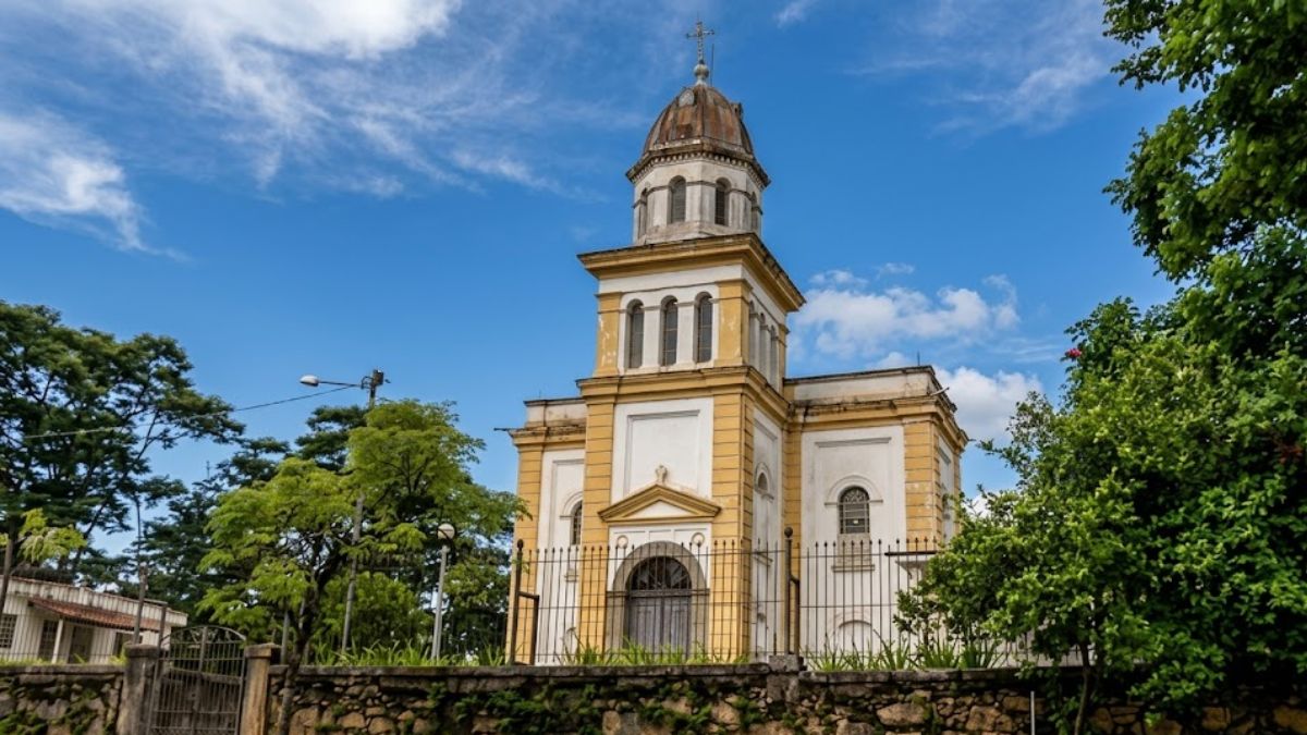 Fotografia realista de uma igreja colonial amarela e branca com torre central sob céu azul ensolarado e cercada por árvores.