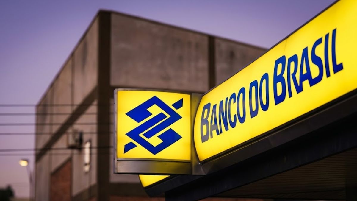 Fotografia em ângulo diagonal da fachada de uma agência do Banco do Brasil com letreiro luminoso amarelo e logotipo azul.