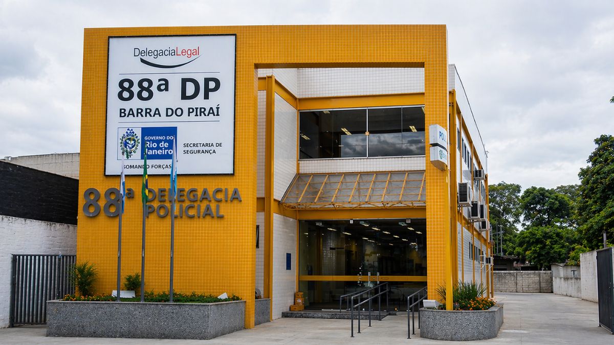 Uma fotografia externa do edifício da 88ª Delegacia de Polícia em Barra do Piraí no Rio de Janeiro com fachada em pastilhas amarelas e um grande letreiro branco. O letreiro exibe os dizeres Delegacia Legal 88ª DP Barra do Piraí Governo do Rio de Janeiro Somando Forças Secretaria de Segurança. Há também três mastros com bandeiras do Brasil e do estado na entrada da unidade policial.