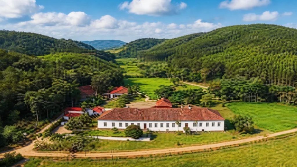 Vista aérea da Fazenda São João da Prosperidade com casarão colonial, áreas verdes extensas e morros ao redor em paisagem rural preservada.