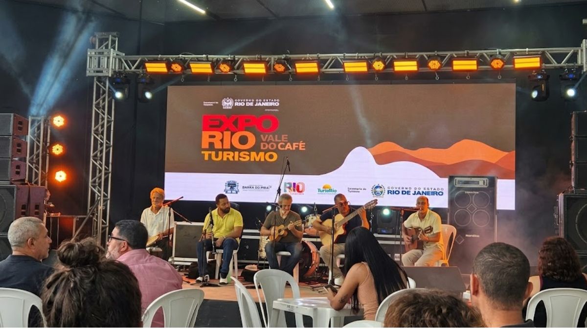 Um grupo de cinco músicos brasileiros, vestindo roupas casuais, toca instrumentos como violão, cavaquinho e percussão em um palco bem iluminado. Um grande telão atrás deles exibe a marca "Expo Rio Vale do Café Turismo" com logotipos oficiais. A platéia está sentada em cadeiras brancas em primeiro plano.