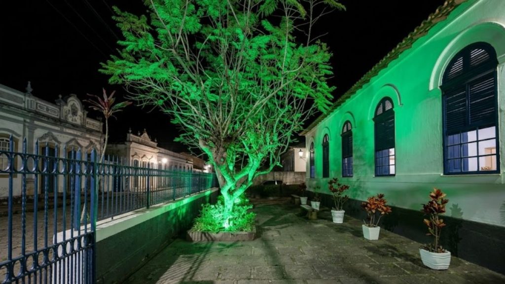 Uma árvore iluminada com luz verde vibrante se destaca em um pátio de pedras ao lado de um casarão histórico branco com janelas azuis durante a noite.