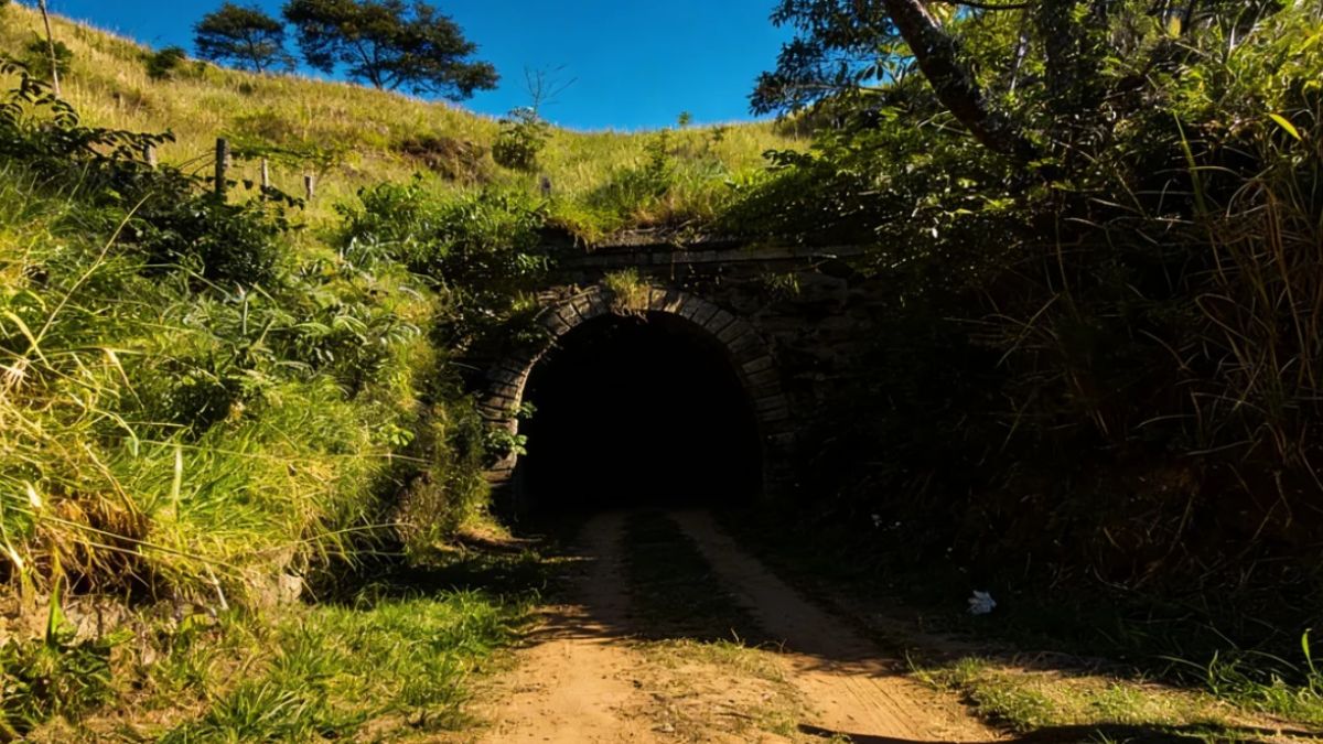 A fotografia captura a saída escura e em arco pleno de um túnel de pedra centenário que leva a uma estrada de terra emoldurada por vegetação densa e uma encosta gramada sob céu azul.