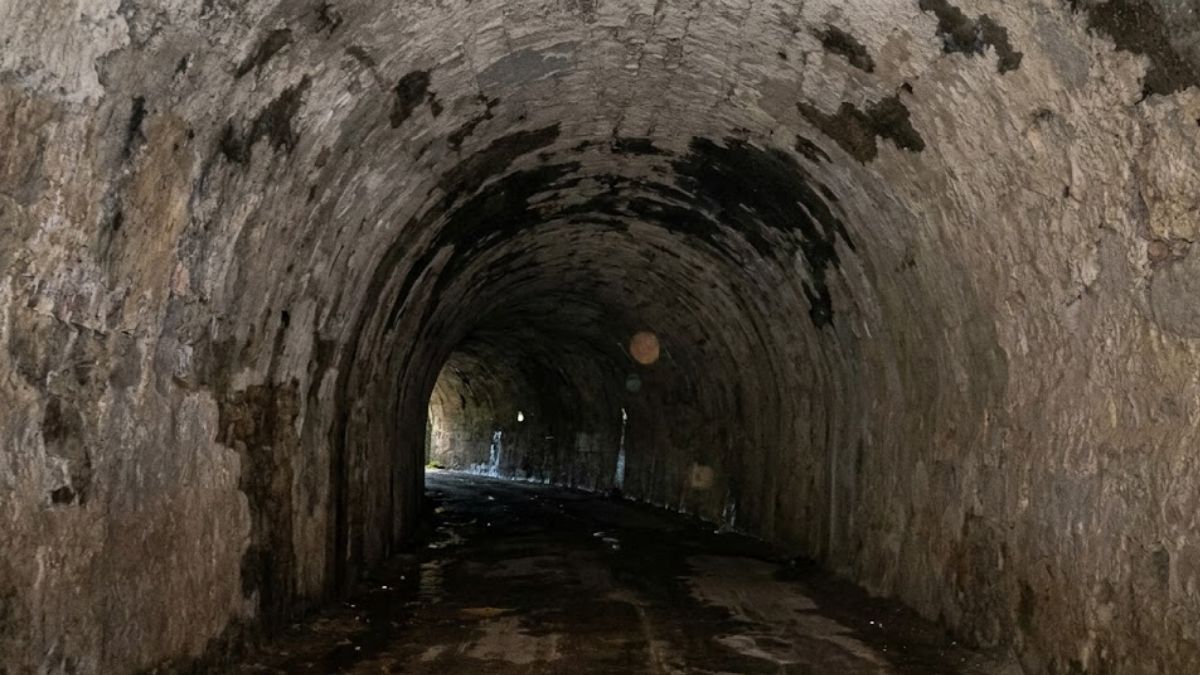 A fotografia captura o interior escuro e curvo de um túnel de pedra centenário com paredes ásperas e úmidas onde o solo irregular e molhado leva a um ponto de luz distante na saída.