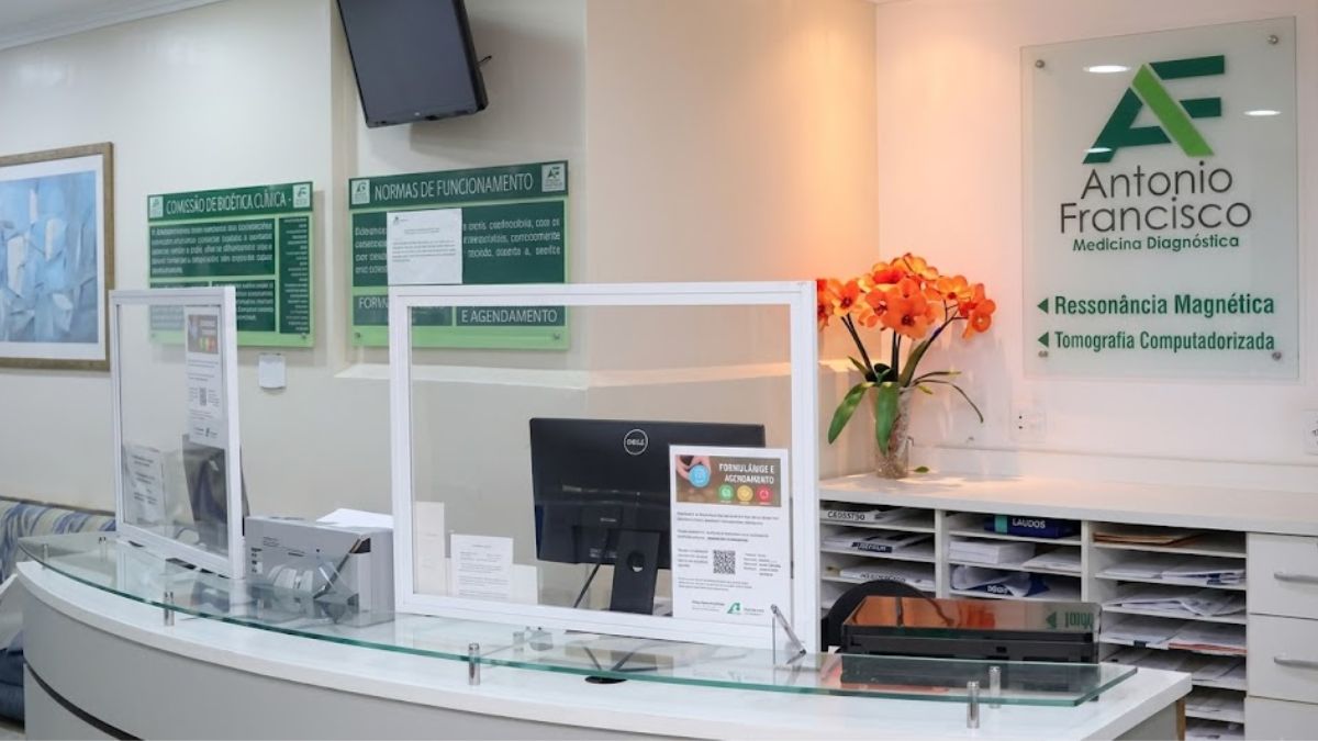 Fotografia da recepção de uma clínica médica com balcão branco curvo, divisórias de vidro, monitores e placa de identificação da marca.