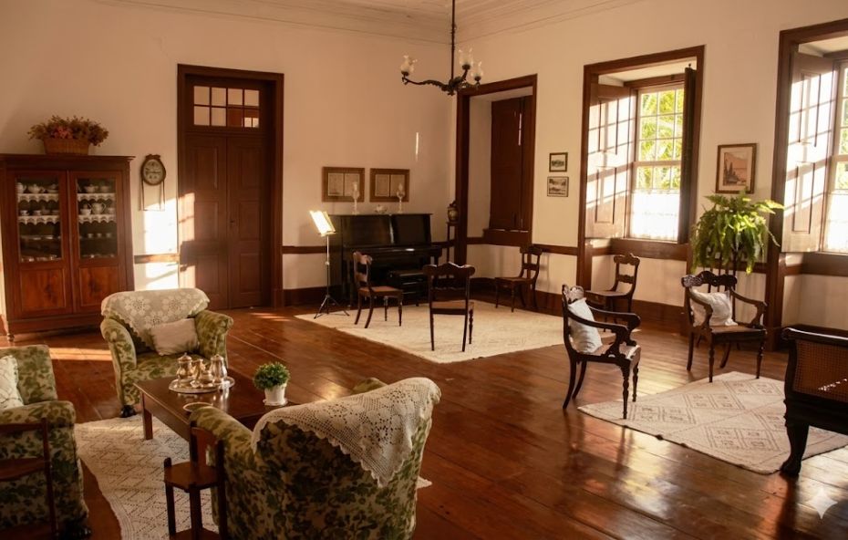 Fotografia realista do interior de um casarão colonial com piso de tábuas largas de madeira escura e paredes brancas. A sala está mobiliada com poltronas estofadas em estampa floral verde, protegidas por rendas brancas nos encostos. Ao fundo, há um piano vertical preto com uma partitura iluminada por um abajur, além de cadeiras de madeira clássicas dispostas sobre tapetes claros. Janelas altas de madeira permitem a entrada da luz solar, que cria sombras longas no chão. Um lustre de metal está pendurado no teto e um armário de madeira com louças completa o ambiente histórico.