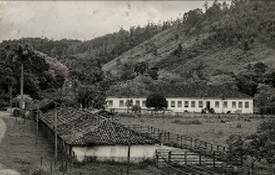 Uma fotografia em preto e branco envelhecida, com marcas de foxing, mostrando uma vista panorâmica de uma fazenda colonial aninhada no Vale do Paraíba. No primeiro plano, à esquerda, há uma longa e rústica estrutura de estábulo com telhado de terracota. Uma cerca de madeira branca delimita Needs e potreiros ao redor de um pequeno jardim central com um arbusto. O casarão principal branco e longo, com dois andares e numerosas janelas, está situado em frente a uma colina verde e íngreme, coberta por pastos e árvores frondosas. No jardim central, figuras humanas em pé podem ser vistas. Outros edifícios de apoio e galpões de beneficiamento de café estão dispostos ao redor. O cenário é rural e histórico, sob luz natural.