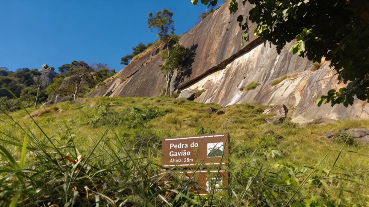 Uma placa marrom de madeira com texto branco indicando Pedra do Gavião Altina 28m está cercada por grama alta em frente a um grande paredão de rocha íngreme sob céu azul límpido.