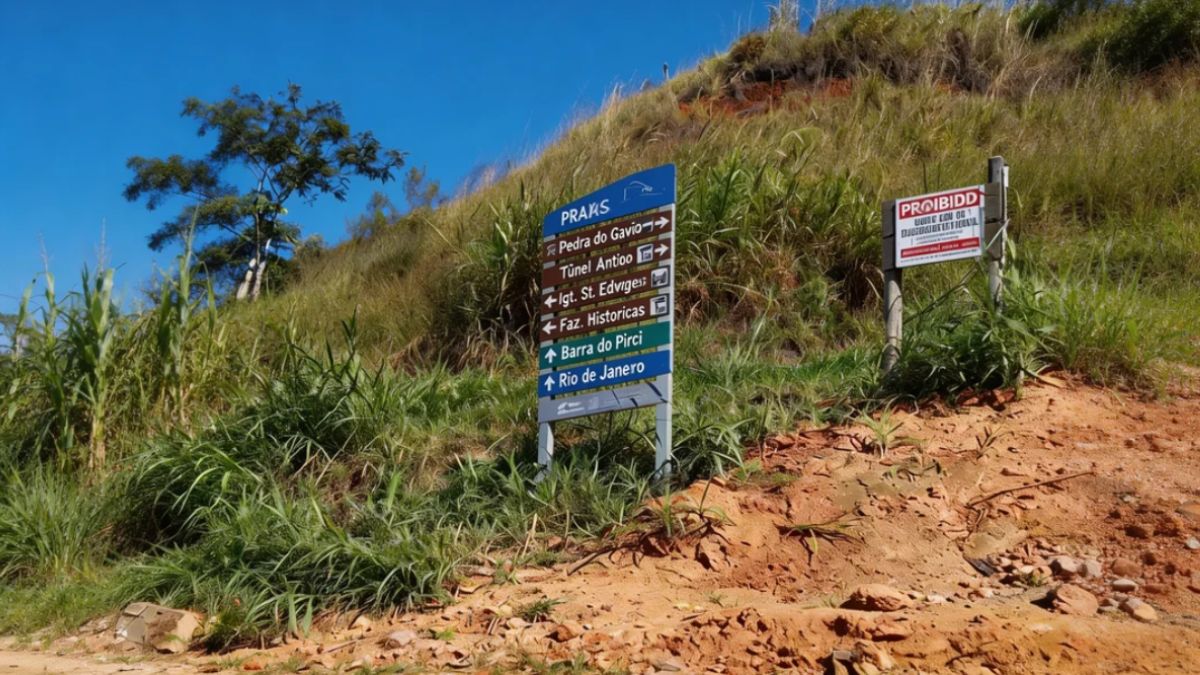 Uma placa azul e branca com setas indicativas para o Túnel Antigo e Pedra do Gavião está posicionada em uma encosta de terra e vegetação sob céu azul.