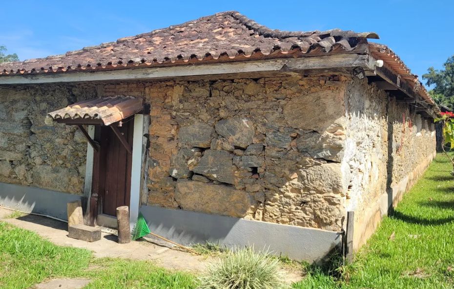 Fotografia em close de uma estrutura rústica de pedra e barro pertencente a uma fazenda histórica. A parede lateral é composta por grandes pedras irregulares assentadas com argamassa de terra, revelando a técnica de construção colonial. O telhado é feito de telhas de barro envelhecidas e moldadas à mão. Há uma pequena porta de madeira escura com um toldo simples de telhas logo acima. Ao lado da porta, um suporte de madeira e uma vassoura de jardim verde estão encostados na base da parede que possui um rodapé pintado de cinza. O gramado verde ao redor e a luz solar direta destacam as texturas das pedras.