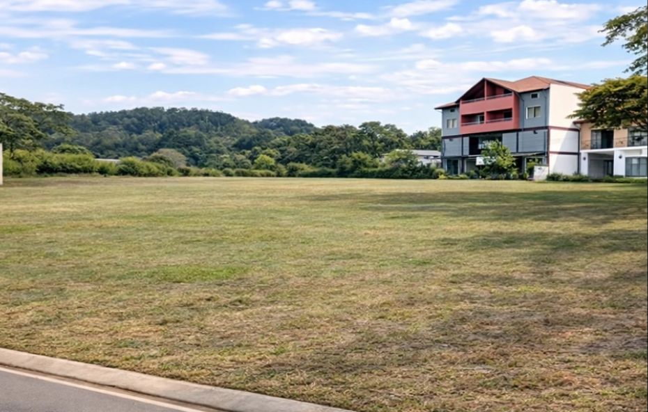 Amplo terreno plano gramado em destaque com um prédio comercial moderno ao fundo sob um céu azul com nuvens esparsas.