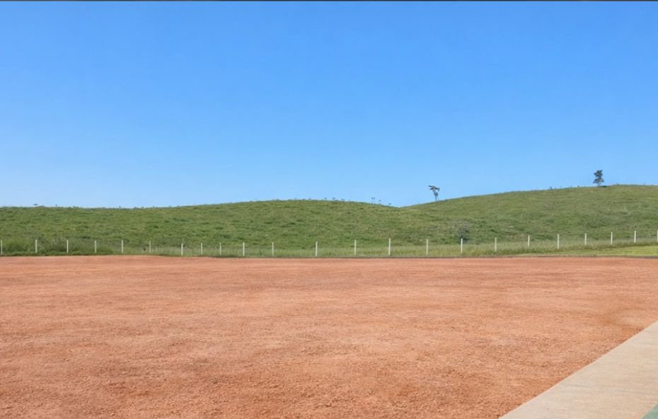 Ampla área plana de terra batida em tons de laranja sob um céu azul limpo com colinas verdes ao fundo.