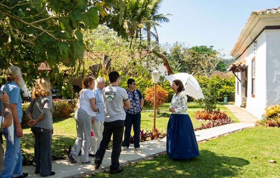 Fotografia colorida capturada em um ângulo médio sob a luz do dia em um jardim gramado. À esquerda, um grupo de seis turistas adultos, homens e mulheres vestidos com roupas casuais contemporâneas, está em pé sobre um caminho de cimento. Eles observam atentamente uma guia turística caracterizada com trajes de época que está posicionada à direita. A guia veste uma blusa branca floral de mangas longas, uma saia longa azul escura e segura um guarda-sol de renda branca. Ao fundo, vê-se a lateral de um casarão colonial branco com janelas de moldura marrom e telhado de barro. A cena é emoldurada por árvores frondosas à esquerda e vegetação tropical rasteira ao longo do caminho, sob um céu claro.