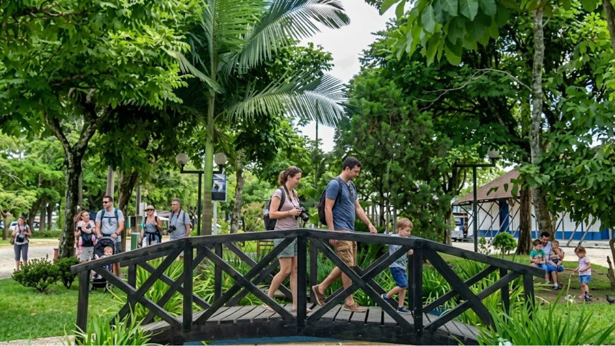 Uma fotografia colorida e realista captada durante o dia em Ipiabas, Barra do Piraí. A imagem mostra diversos turistas atravessando uma ponte de madeira em uma praça arborizada e verde, com uma família em destaque e outras pessoas ao fundo.