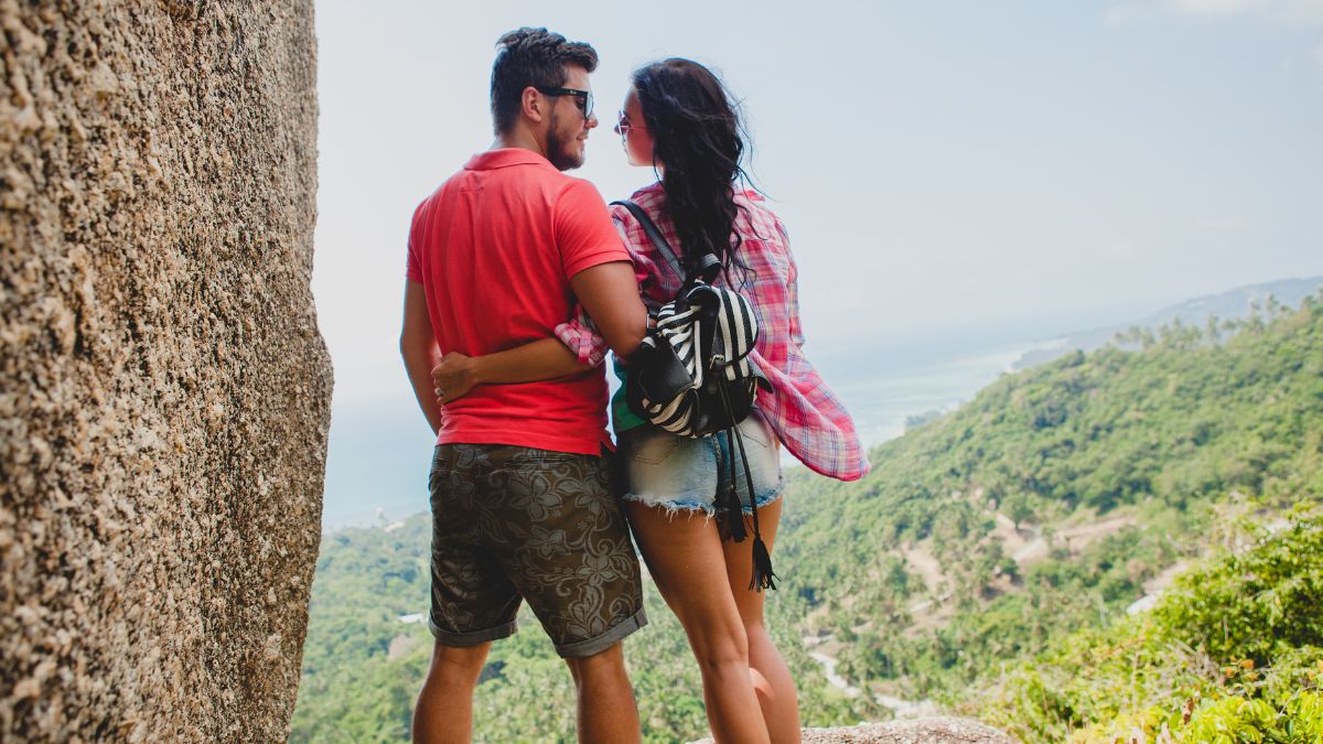 Um casal jovem, visto de costas, se abraça em um mirante rochoso enquanto admira a vista panorâmica de montanhas verdes e o mar ao longe, em um dia ensolarado.