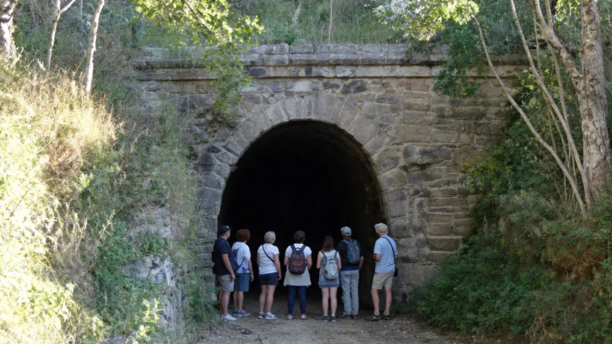 Sete turistas com mochilas e trajes de caminhada aparecem de costas observando a entrada em arco pleno do Túnel Velho de Ipiabas que está encravada em uma parede de pedras centenárias cercada por mata verde densa.