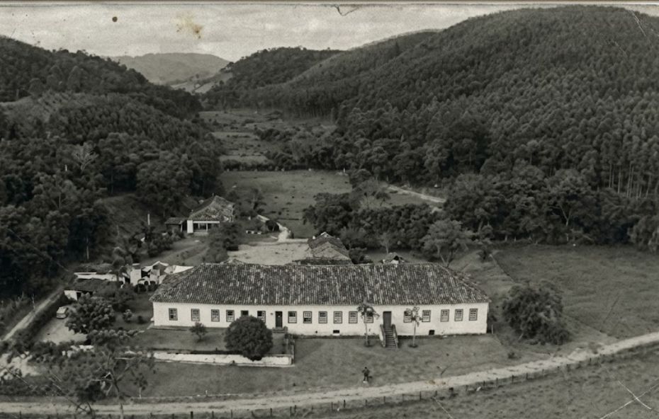 Fotografia aérea em preto e branco de meados do século vinte mostrando a sede de uma grande fazenda colonial de café no Vale do Paraíba. O complexo inclui um casarão principal branco e longo com telhado de terracota, localizado no pé de uma colina íngreme coberta por floresta nativa densa e potreiros gramados. Um pátio de serviço central e de trabalho por trás da residência exibe várias carroças puxadas por cavalos e figuras humanas organizadas. Outras construções de apoio, incluindo galpões de beneficiamento de café, tulhas, e estrebarias, estão dispostas ao redor, juntamente com cercas e potreiros gramados com gado. A paisagem rural estende-se ao fundo sob luz natural.