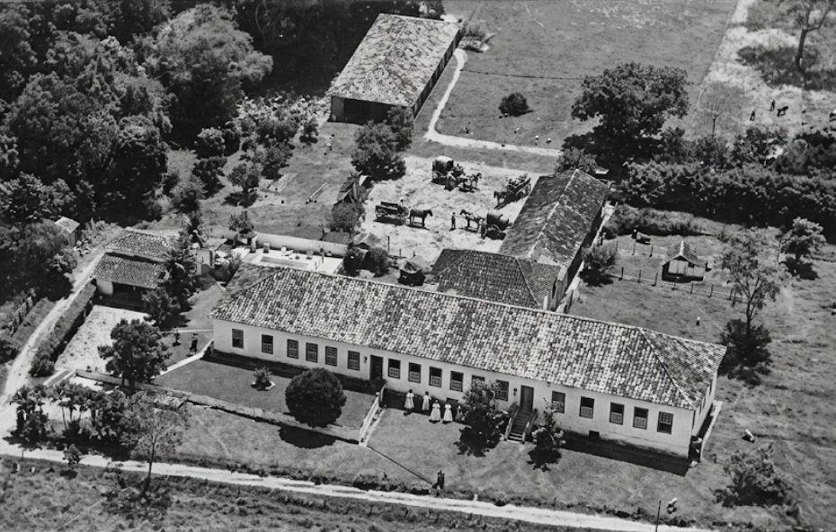 Fotografia aérea em preto e branco de meados do século vinte mostrando um grande complexo de fazenda colonial. No primeiro plano, uma longa casa de fazenda branca de um andar com telhado de terracota está situada em frente a um jardim gramado. A área de pátio central e de trabalho por trás da casa principal está repleta de várias carroças puxadas por cavalos e figuras humanas. Outros edifícios de apoio, galpões e potreiros gramados, cercados por vegetação densa ao redor e floresta ao fundo, completam o cenário. O sol brilha forte.