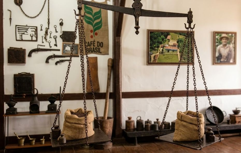 Fotografia aérea histórica em preto e branco mostrando uma visão panorâmica e detalhada de uma grande fazenda colonial de café. No primeiro plano, o casarão principal longo e branco com telhado de terracota. Imediatamente atrás da sede, estende-se um vasto pátio operacional composto por um grande terreiro de secagem de café, calçado com pedras brutas e com caminhos de terra. Várias figuras humanas e animais de carga, incluindo carroças puxadas por cavalos e mulas, estão dispostos no terreiro, sugerindo a atividade produtiva. Ao redor do terreiro, várias construções de apoio estão posicionadas: tulhas adjacentes de madeira com ventilação, galpões de beneficiamento de café, estábulos, e moradias de trabalhadores. A paisagem rural estende-se além do complexo, com pastagens gramadas, cercas, potreiros, e uma floresta nativa densa cobrindo as colinas ao fundo sob luz natural.