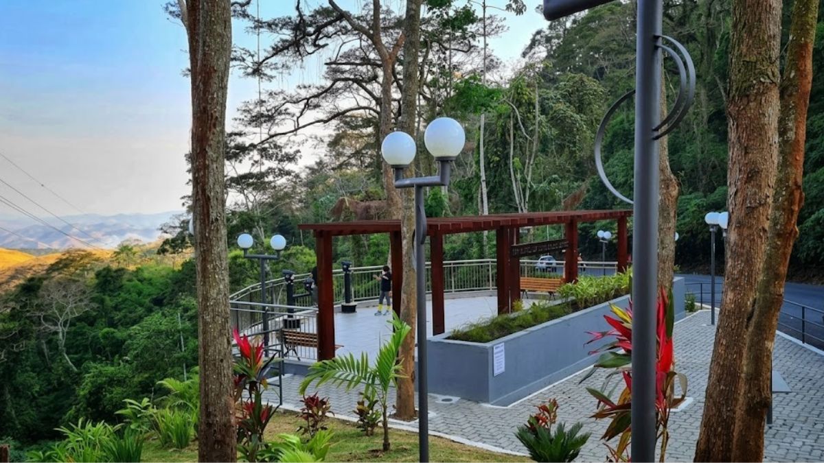 Fotografia colorida do Mirante de Ipiabas, no Rio de Janeiro. A imagem mostra a plataforma de observação pavimentada com piso intertravado, rodeada por árvores nativas e um jardim com plantas vermelhas e verdes em primeiro plano. O mirante possui postes de luz modernos com globos brancos e uma pérgula de madeira coberta por telhas vermelhas. Um visitante está na plataforma, observando a vista. Ao fundo, uma vasta paisagem de montanhas cobertas por vegetação densa se estende sob um céu claro. Uma estrada e um carro são visíveis ao lado do mirante.