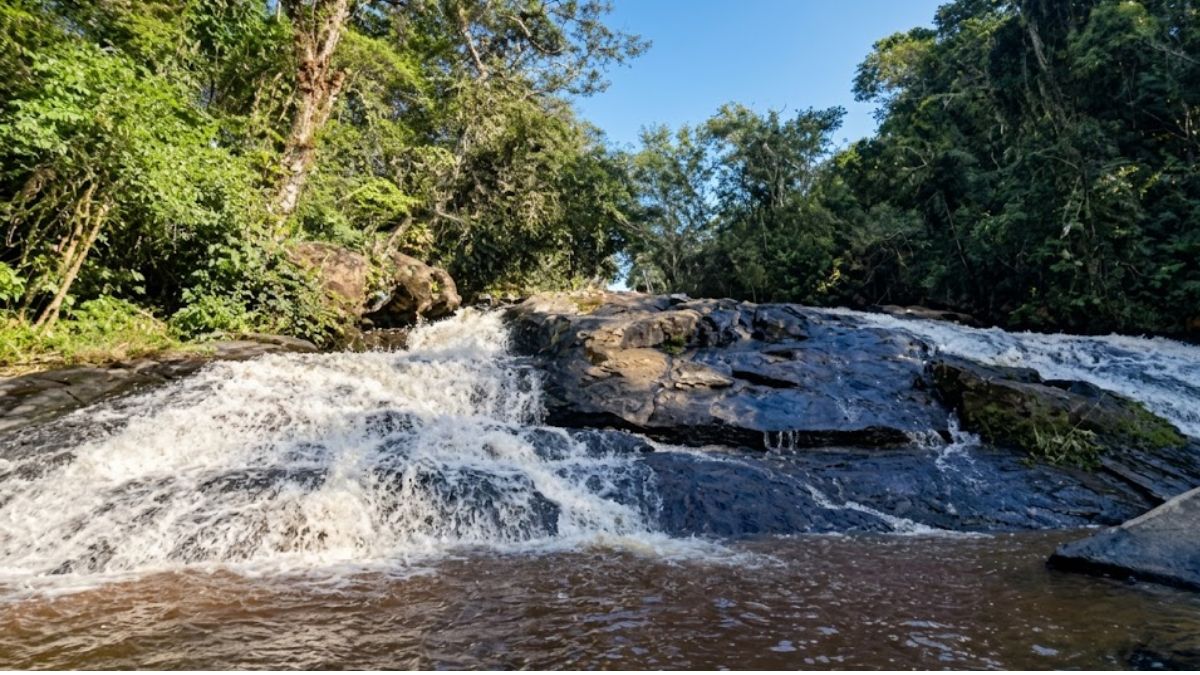 Uma fotografia realista em close-up da Cachoeira de Ipiabas, em Barra do Piraí, Rio de Janeiro. A imagem captura a água correndo sobre rochas escuras e desgastadas, caindo em um lago de cor avermelhada. No topo da cachoeira, há uma densa floresta verde de Mata Atlântica sob um céu azul claro.