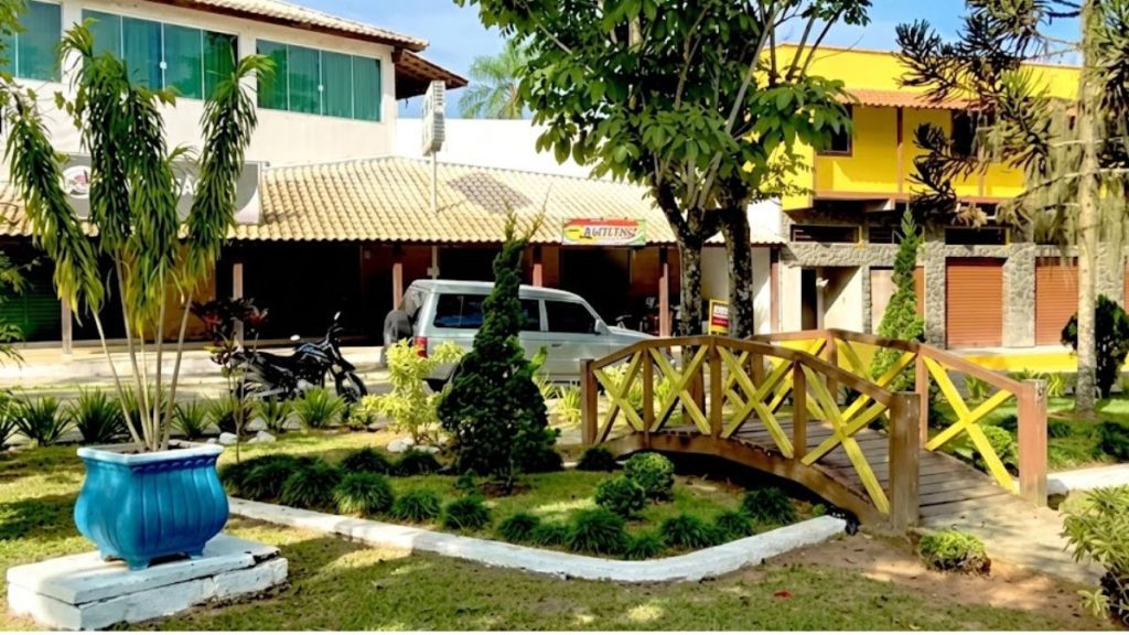 Uma fotografia realista em 4K e alta definição mostrando um jardim urbano bem cuidado com uma ponte de madeira e edifícios comerciais em Ipiabas, Rio de Janeiro. A ponte de madeira arqueada com detalhes amarelos atravessa o gramado, e à esquerda, um grande vaso de plantas azul brilhante está sobre uma base branca. Ao fundo, um SUV prateado e uma motocicleta preta estão estacionados em frente a prédios comerciais com toldos e janelas verdes, com árvores altas e vegetação densa ao redor.