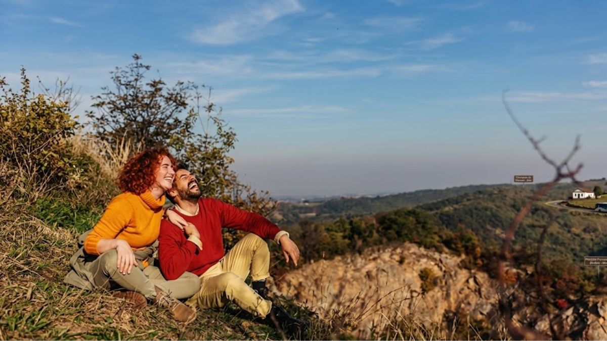 Um casal sorridente, com a mulher de cabelo ruivo e o homem de suéter vermelho, está sentado no topo de um morro rochoso e gramado. Eles olham para cima e para a direita, dando risada. Ao fundo, uma vasta paisagem de colinas verdejantes e vales se estende sob um céu azul claro com nuvens leves. Uma pequena construção e uma estrada são visíveis em um cume distante.