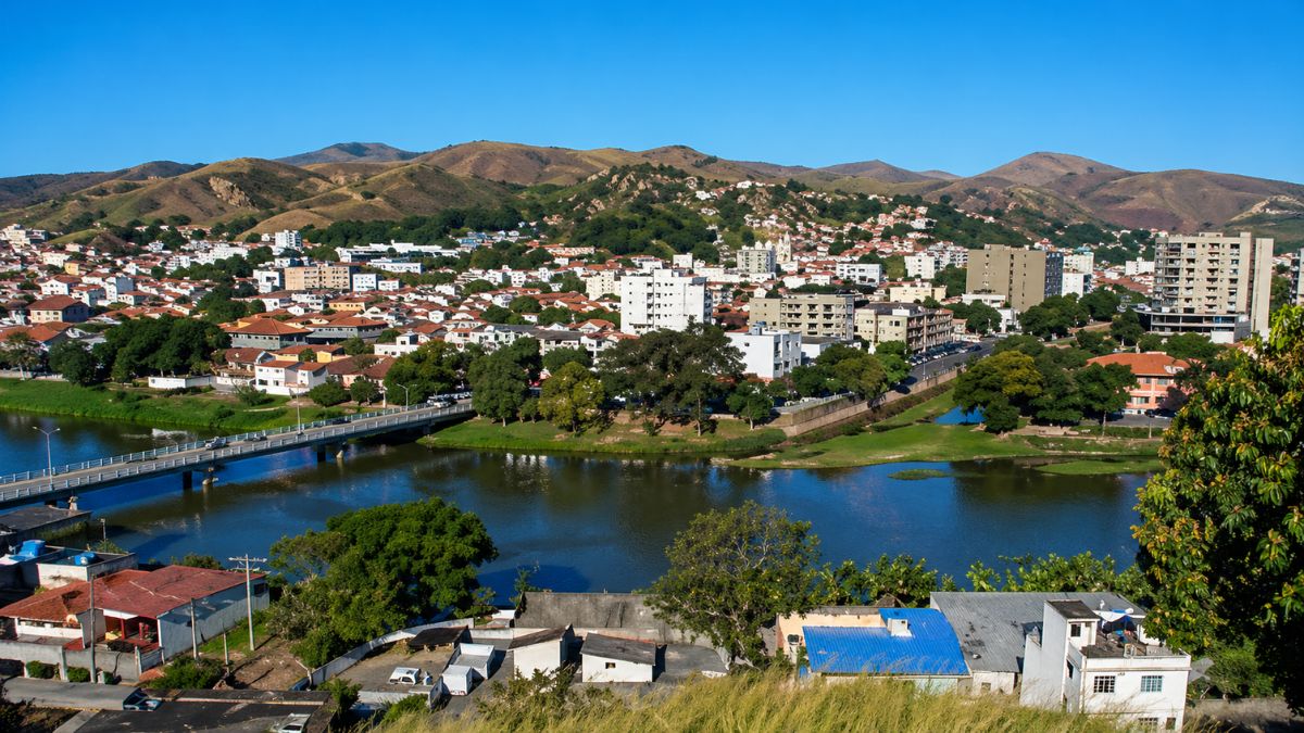 Fotografia aérea de Barra do Piraí mostrando o Rio Paraíba do Sul, uma ponte rodoviária, edifícios brancos e montanhas ao fundo.