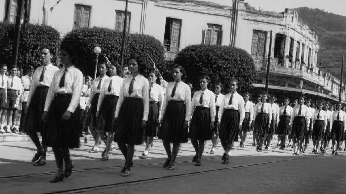 Fotografia antiga em preto e branco mostrando uma fileira de jovens estudantes marchando uniformizadas em uma rua com trilhos.